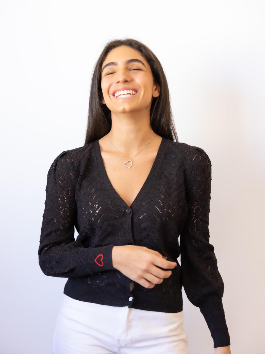 Black alpaca cardigan featuring a delicate open-knit pattern and a small red embroidered heart on the cuff, shown here on a model against a plain background.