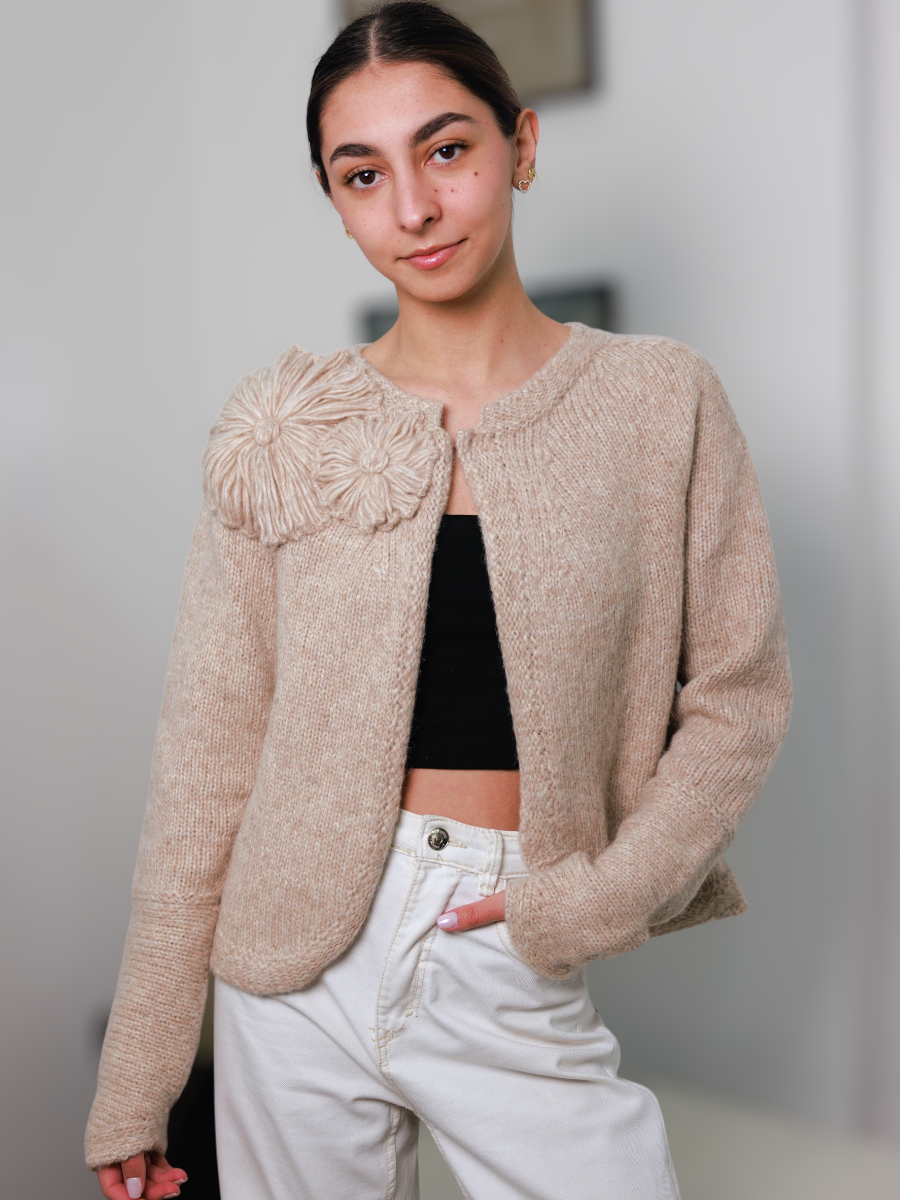 Woman wearing a beige textured cardigan with a floral embellishment, black top, and white pants.
