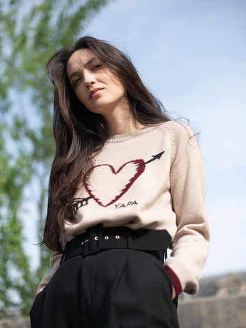 beige alpaca sweater featuring a heart with an arrow design on the chest on model in city street
