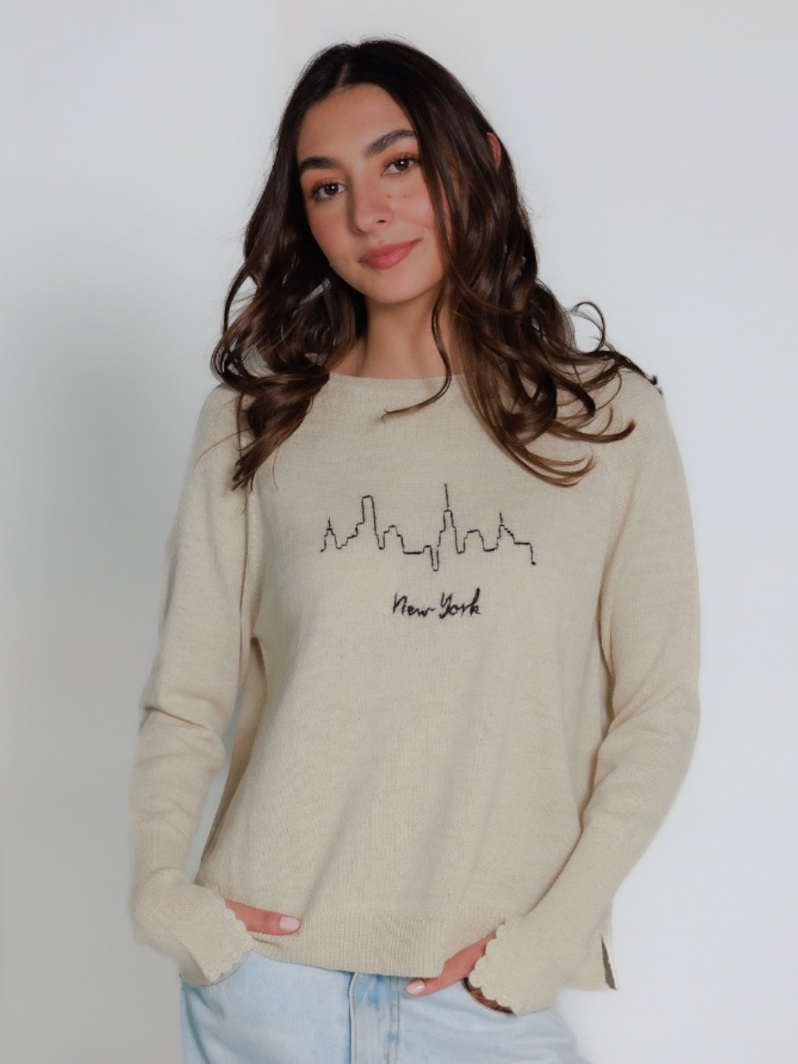 Woman wearing a beige sweater with New York and skyline design on a plain background