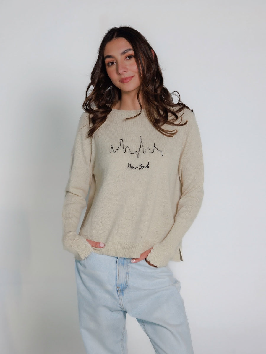 Model wearing a white hand embroidered sweater with 'New York' text and skyline on a plain background
