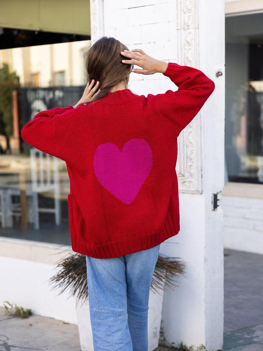 Red alpaca cardigan featuring an oversized pink heart motif on the back, shown here on a model in an outdoor urban setting.