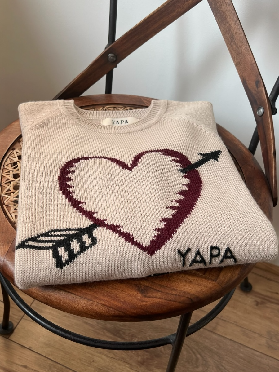 beige alpaca sweater featuring a heart with an arrow design on the chest folded on a chair