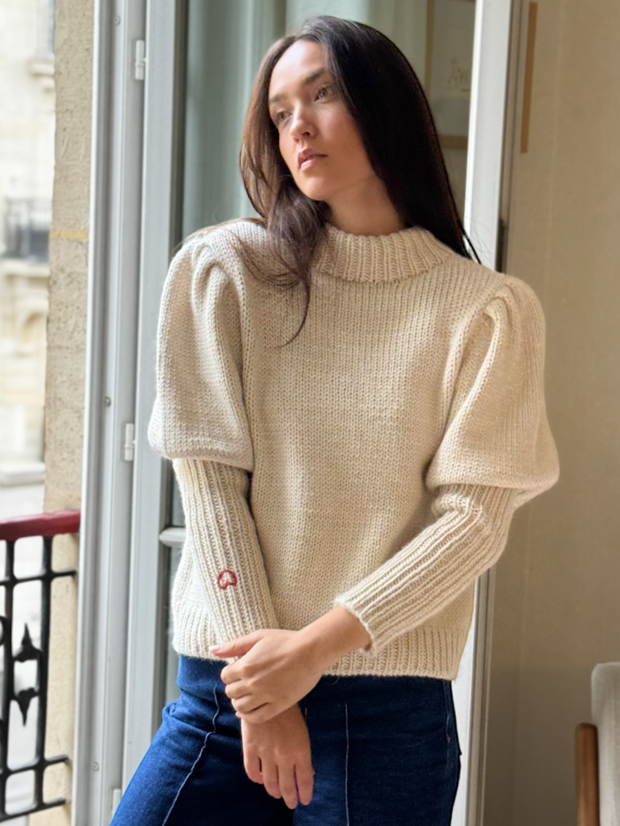 White alpaca cardigan featuring puffed sleeves, a ribbed high neckline, and a small red embroidered heart on the cuff, shown here on a model in an indoor setting.