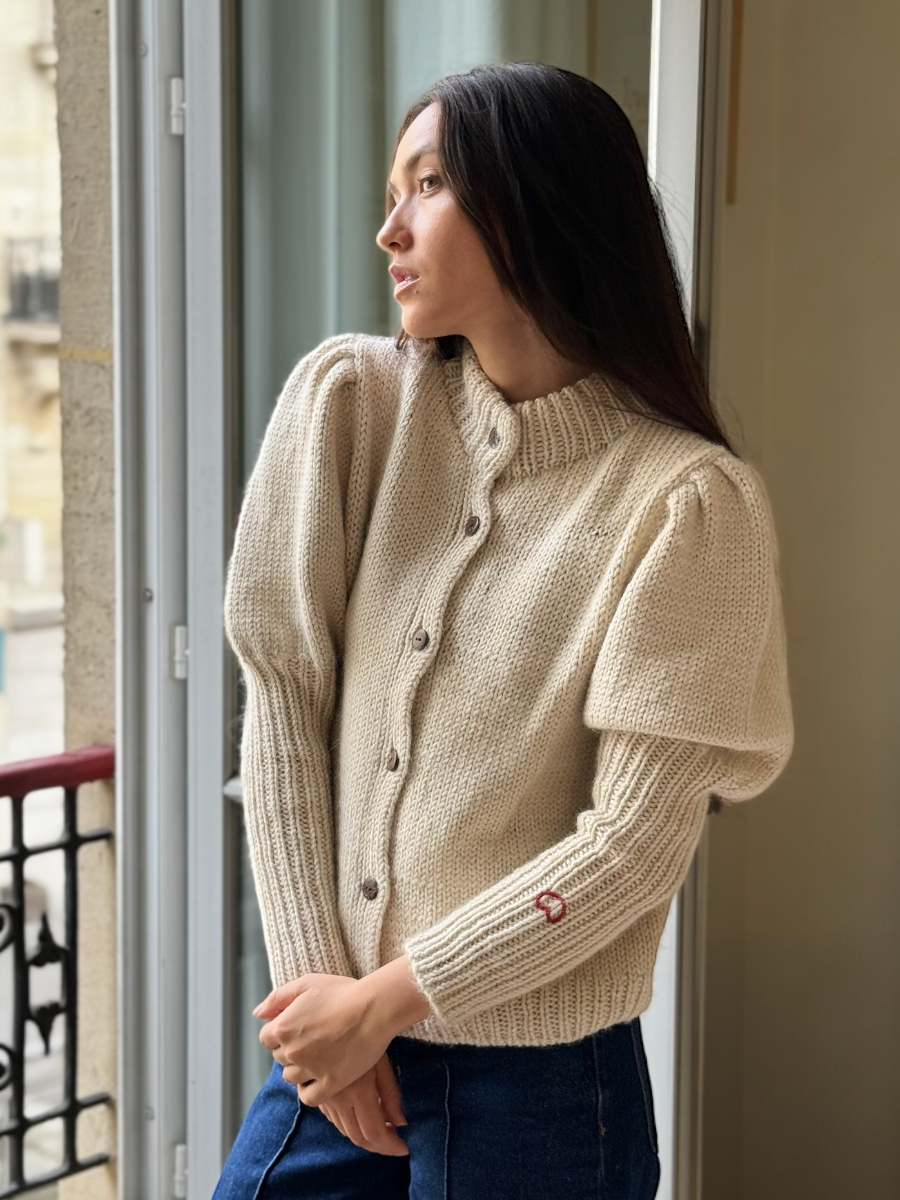 White alpacacardigan featuring puffed sleeves, a ribbed high neckline, and a small red embroidered heart on the cuff, shown here on a model in an indoor setting.