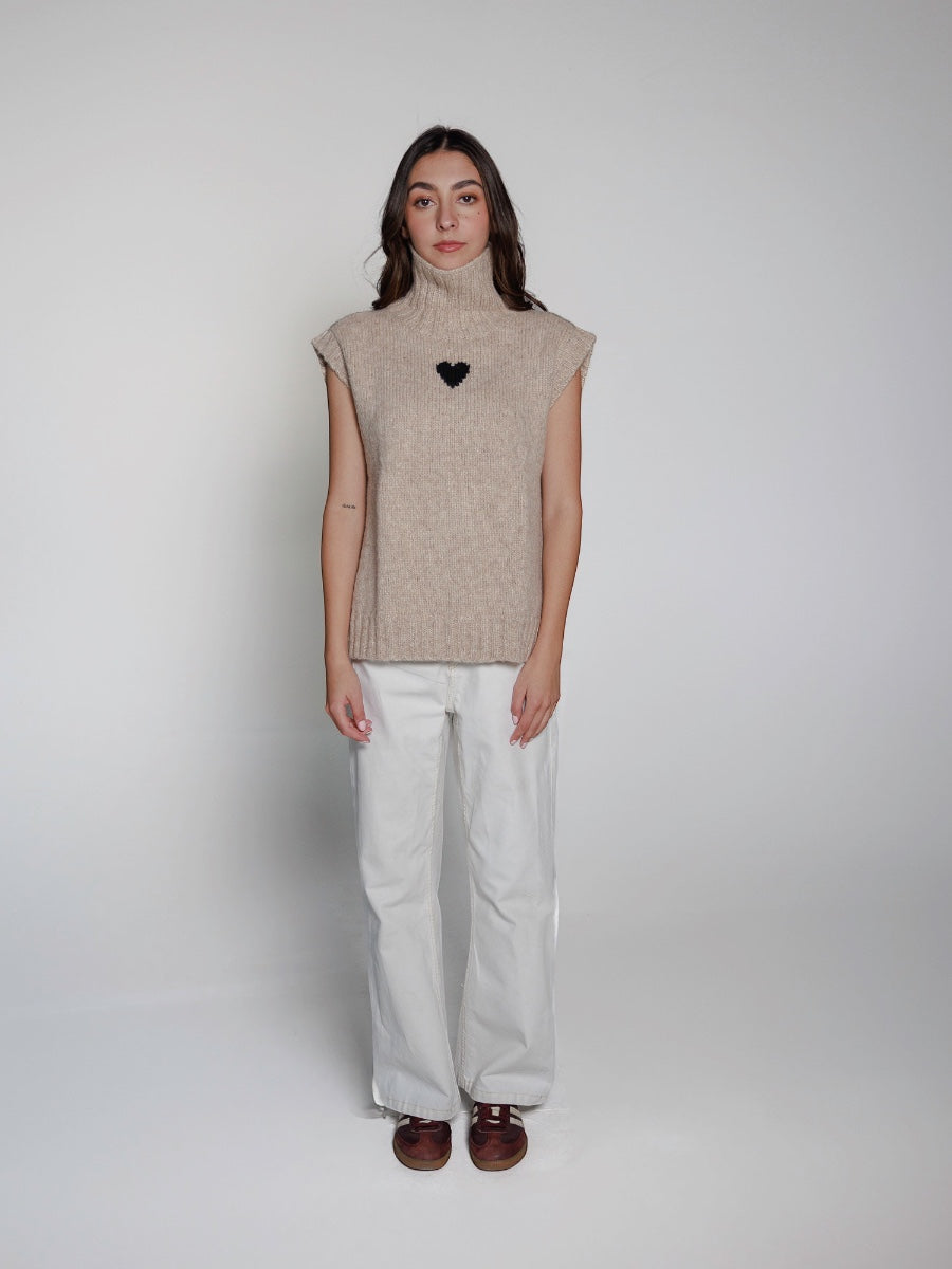 model wearing a beige sleeveless alpaca sweater with a black heart design and white pants on a plain background