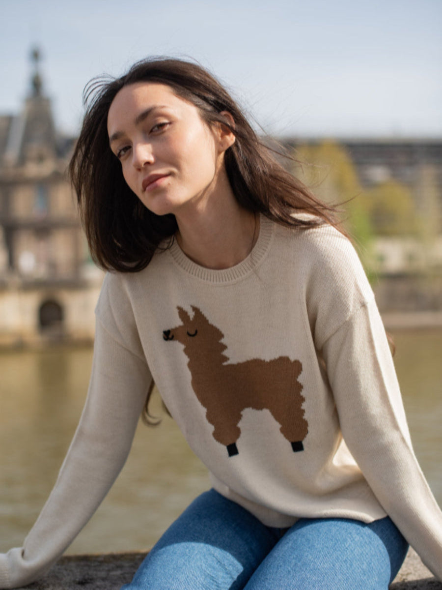 beige alpaca sweater with a brown llama motif, worn by a model styled with blue jeans with a city and river background.