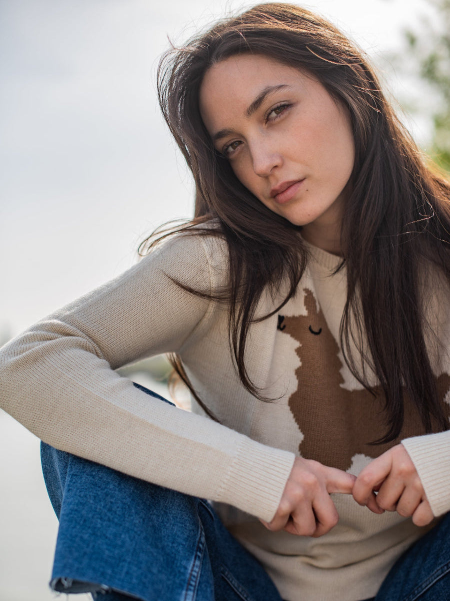 beige alpaca sweater with a brown llama motif, worn by a model styled with blue jeans with a city and river background.