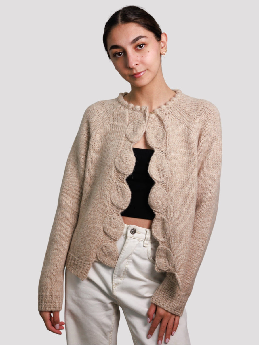 beige llama cardigan with circular knitted details along the front edges, worn by a model with white pants and a black top against a plain background.