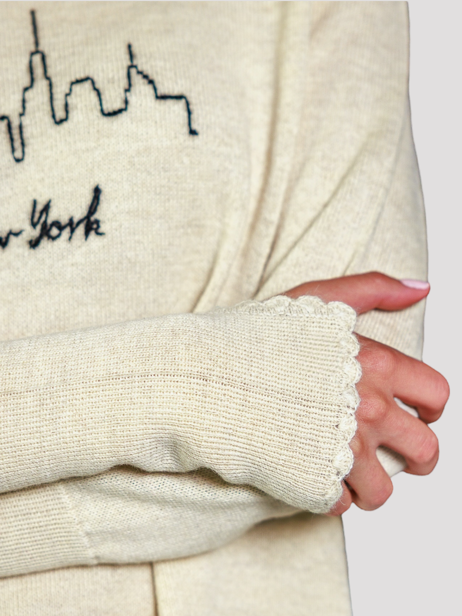 Close-up of a cream-colored sweater with embroidered New York skyline design, held by a person.