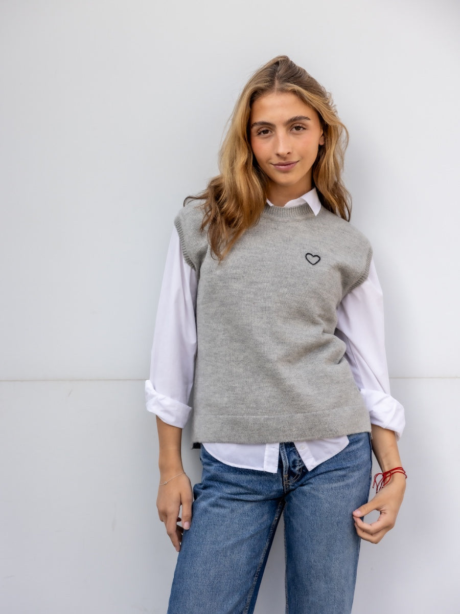 Woman wearing a gray sleeveless sweater over a white shirt with blue jeans against a light background
