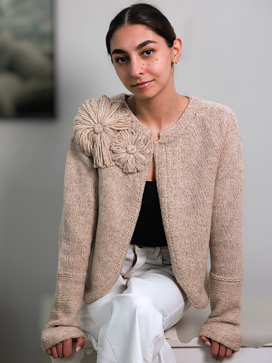 Woman wearing a beige textured cardigan with a floral embellishment, black top, and white pants.