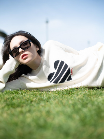 white alpaca knitted sweater with a black striped heart design, worn by a model lying on the grass