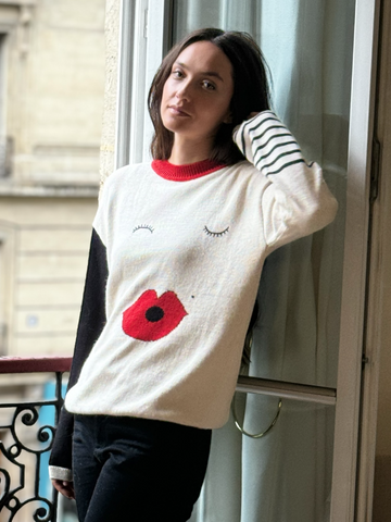 Playful white alpaca knitted sweater featuring a red neckline with a face design with closed eyes, bold red lips, and contrasting sleeves, one black and the other with stripped lines, shown on a model by a window.