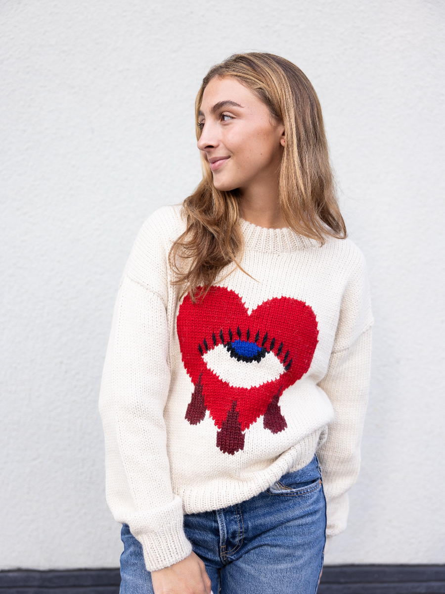 white alpaca sweater featuring a bold red heart motif with a blue eye and tear details on the front, shown here on a model against a plain background.