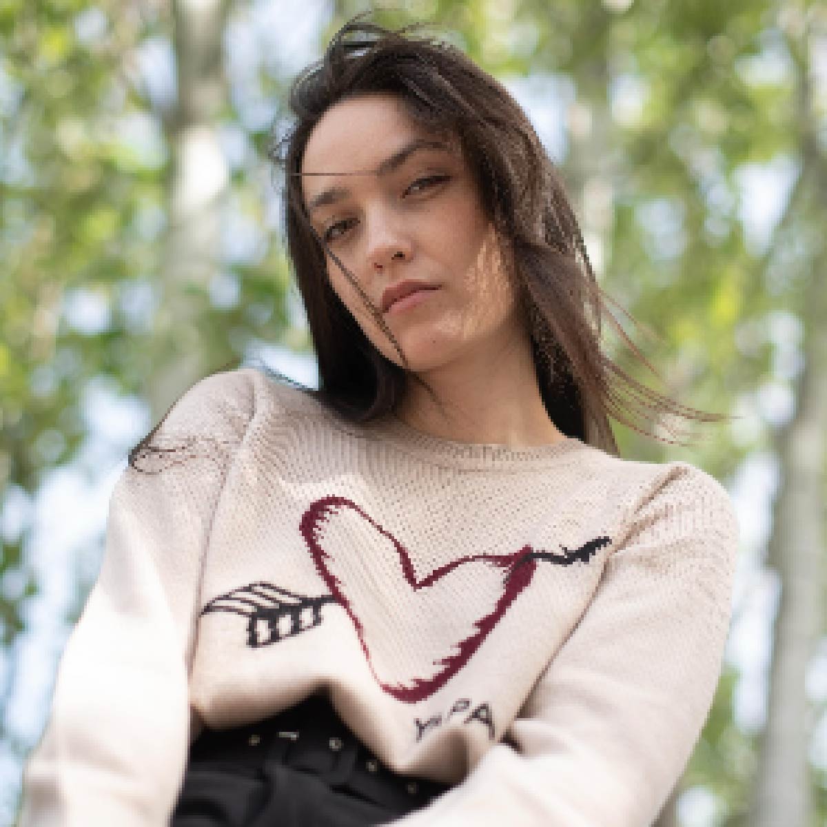 beige alpaca sweater featuring a heart with an arrow design on the chest on model with trees on the background 