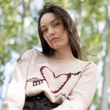 beige alpaca sweater featuring a heart with an arrow design on the chest on model with trees on the background 