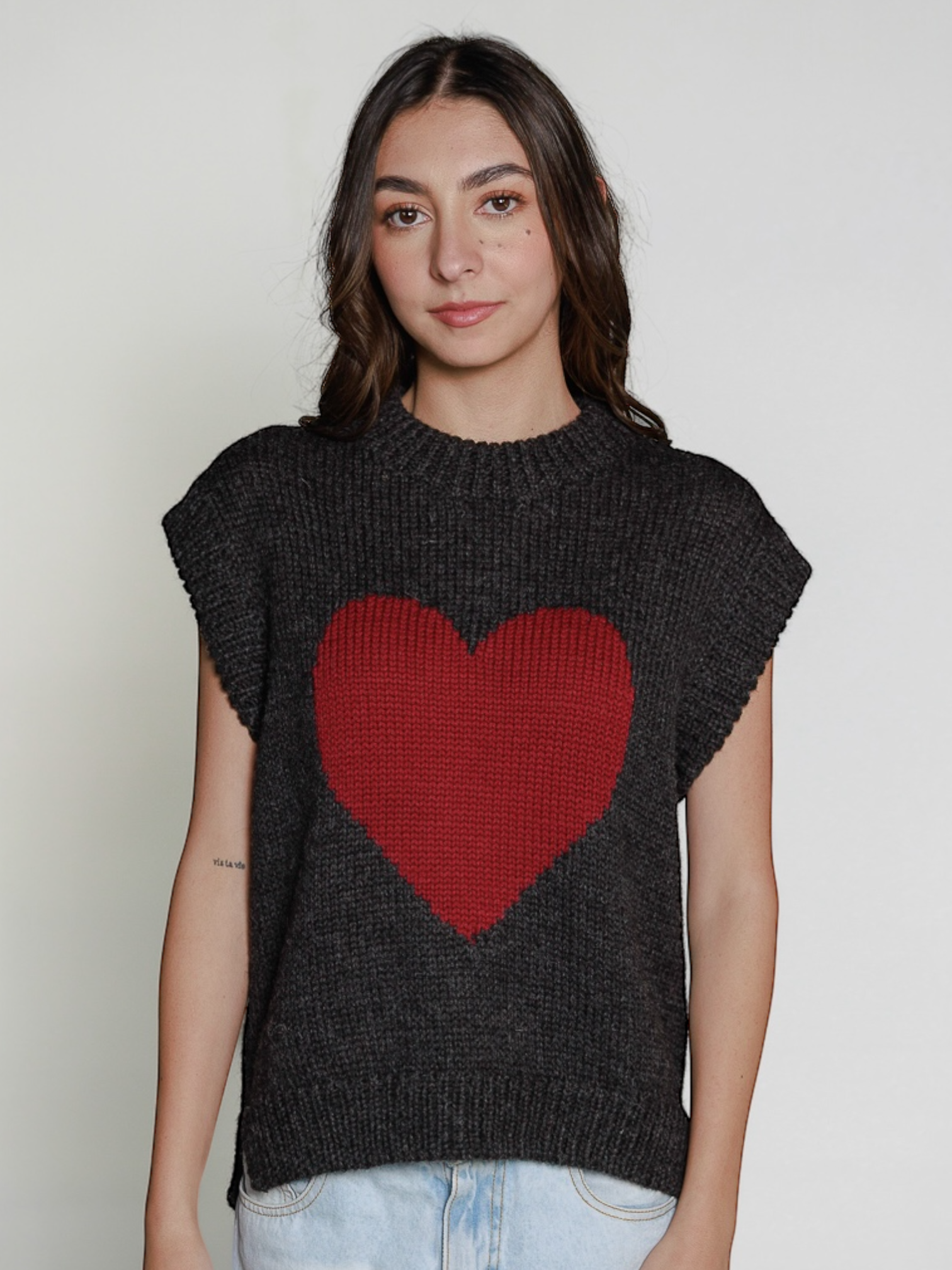 Woman wearing a dark grey sleeveless sweater vest with a red heart design against a plain background