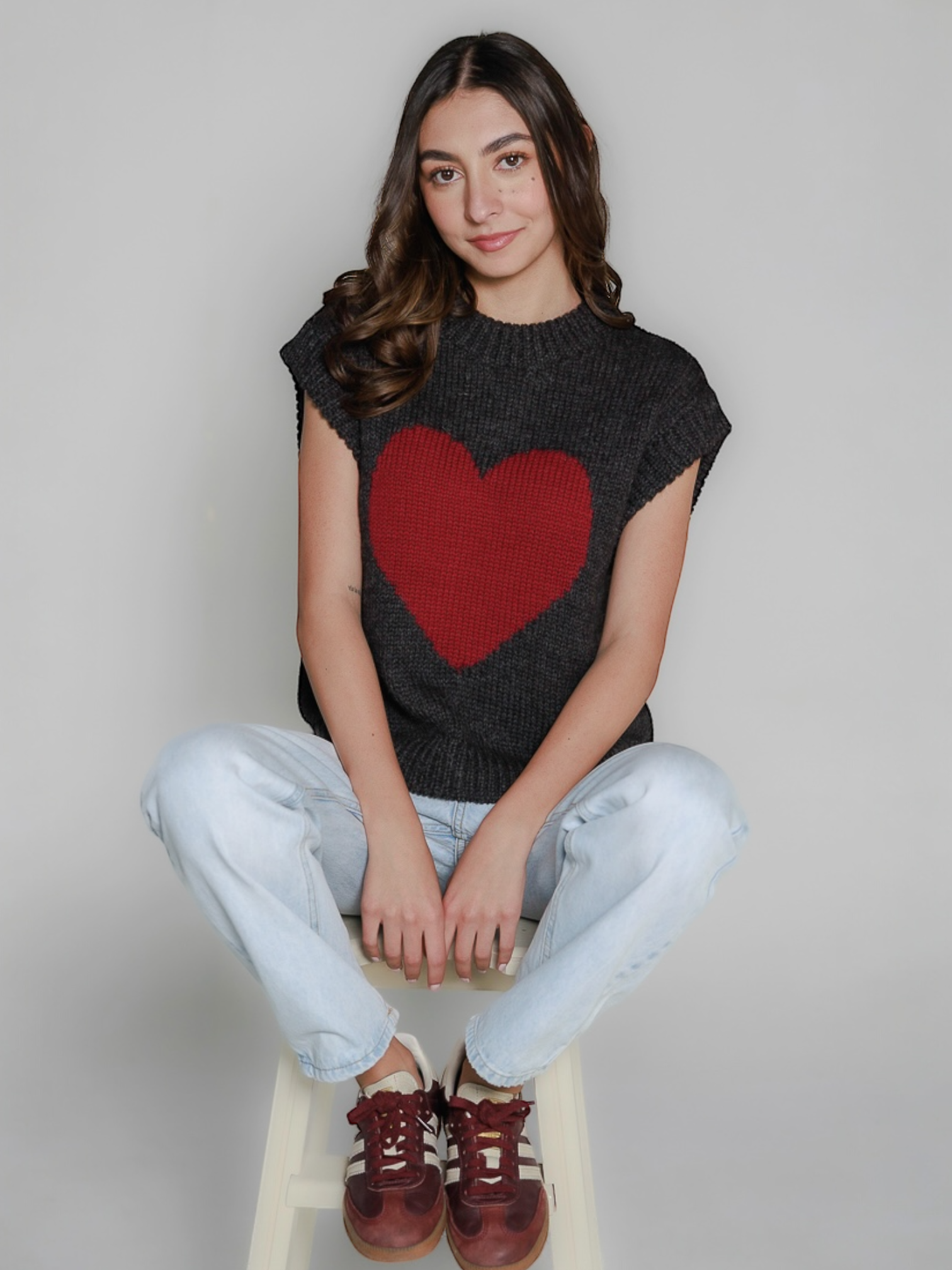 Woman wearing a dark gray sleeveless sweater with a red heart design, sitting on a white stool against a plain background.