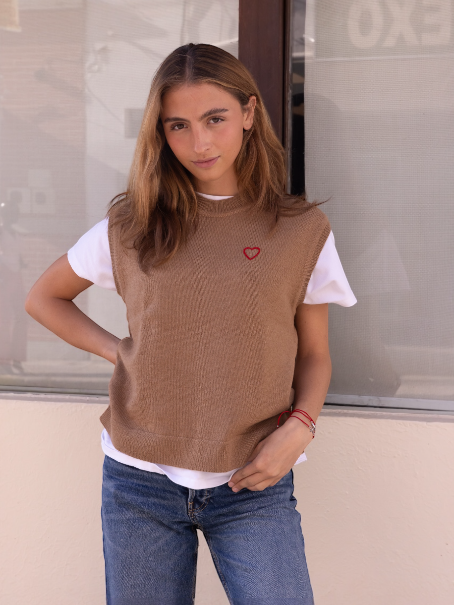 Alpaca sweater vest in a camel color with a red heart detail on model with a wall background
