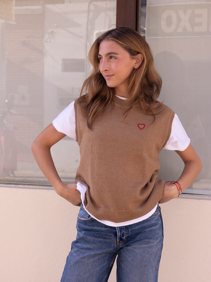 Alpaca sweater vest in a camel color with a red heart detail on model with a wall background