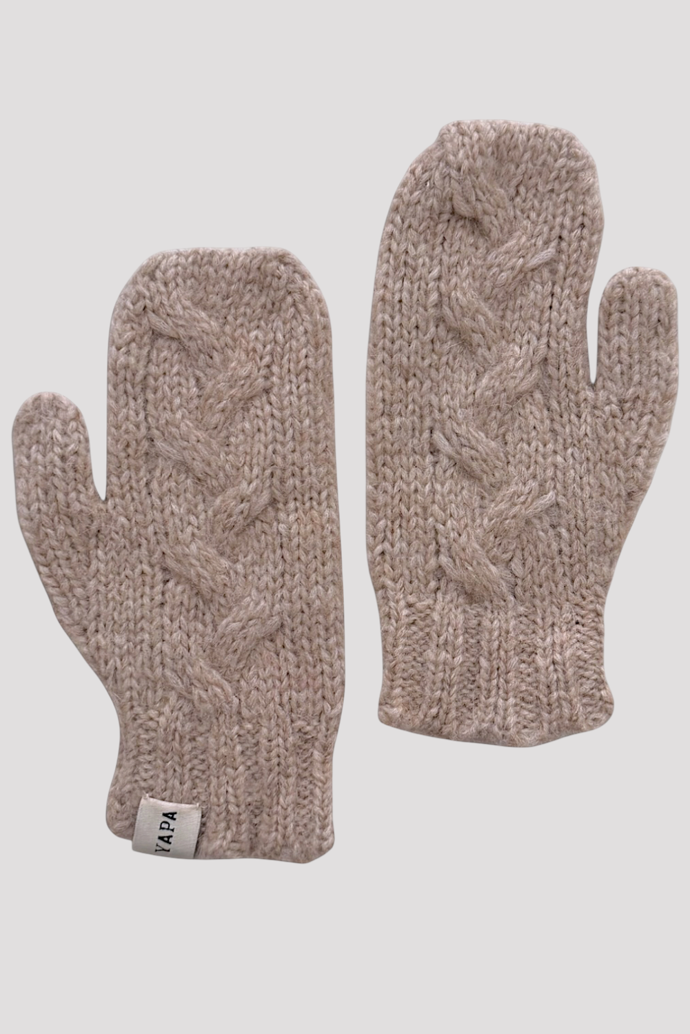 A pair of chunky cable-knit winter mittens in a heathered taupe-beige color, displayed against a solid light gray background. One mitten is shown palm-side and the other is shown finger-side, with a small white brand tag visible on the cuff.