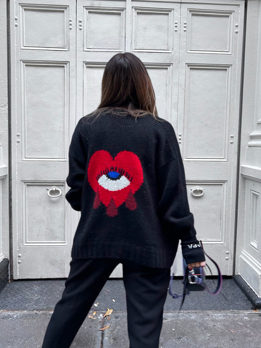 Black alpaca cardigan featuring a bold red heart motif with a blue eye and tear details on the back, shown here on a model in an urban outdoor setting.