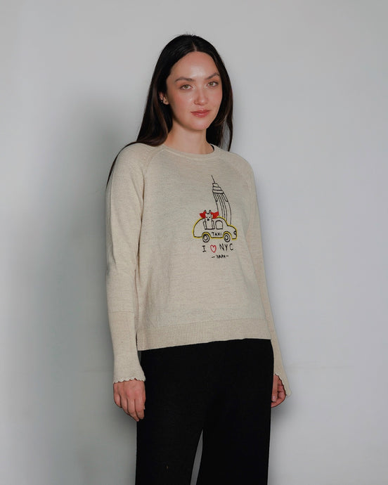Woman wearing a beige sweater with a hand embroidered sweater with a empire estate and llama in a  taxi design against a plain background