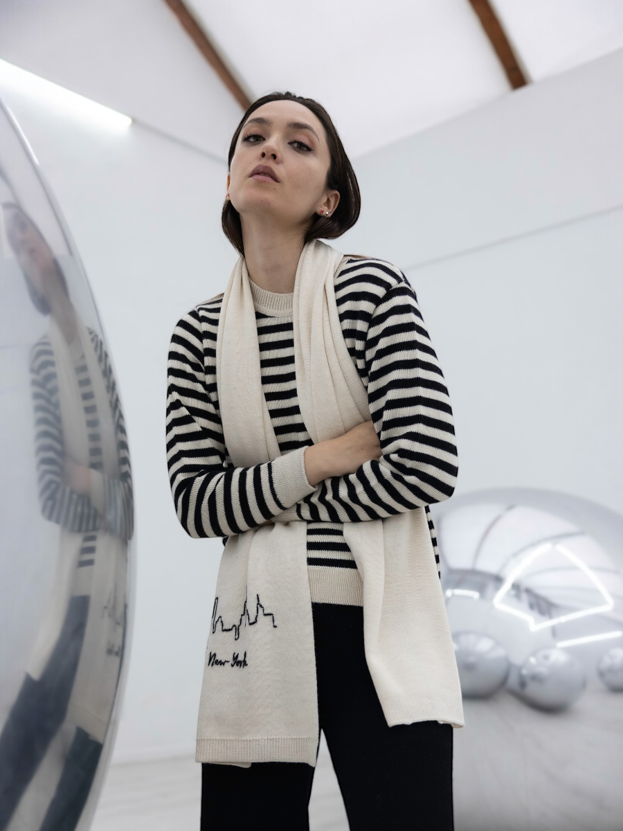 Woman wearing a striped sweater and a long cream scarf with 'New York' print in a modern indoor setting.