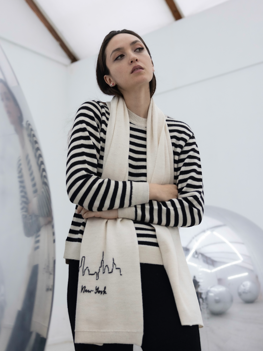White alpaca scarf featuring a hand-embroidered black New York City skyline motif on the front, shown here on a model in an art gallery.