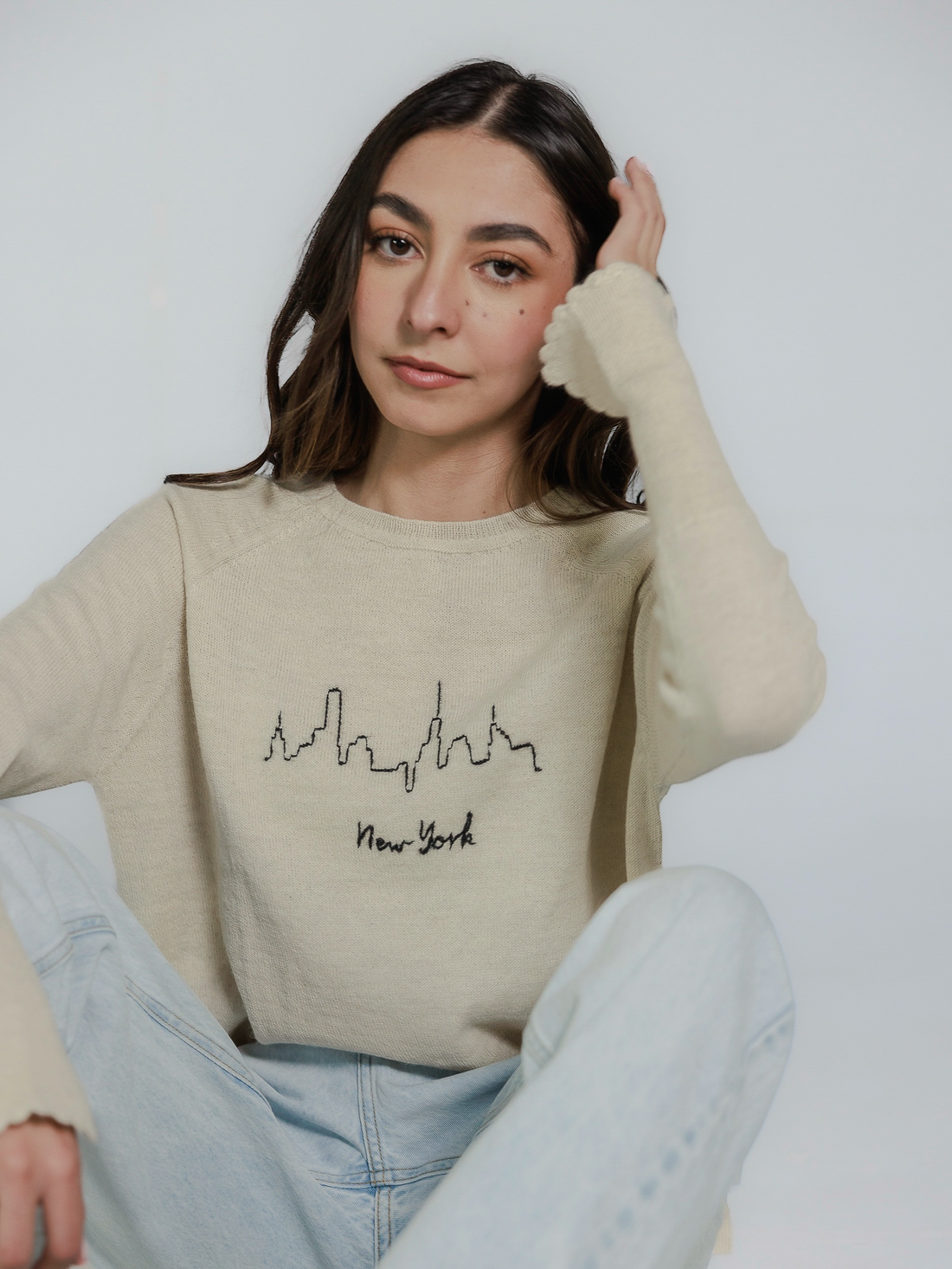 Woman wearing a beige sweater with New York and skyline design on a plain background