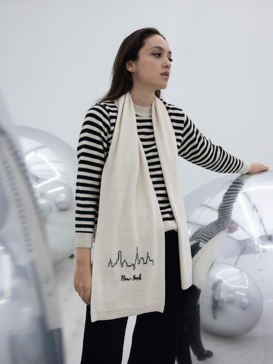 White alpaca scarf featuring a hand-embroidered black New York City skyline motif on the front, shown here on a model in an art gallery.