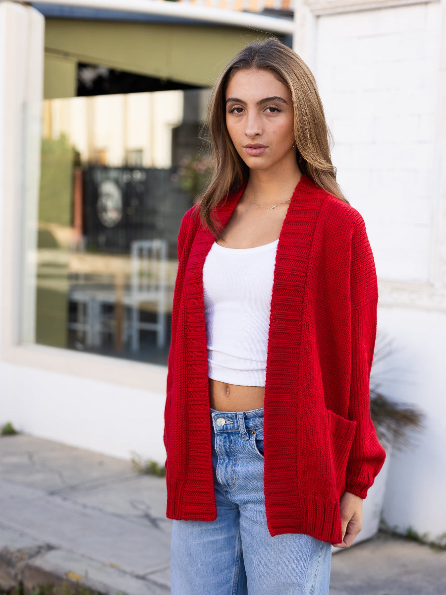 Red alpaca cardigan featuring an oversized pink heart motif on the back, shown here on a model in an outdoor urban setting.