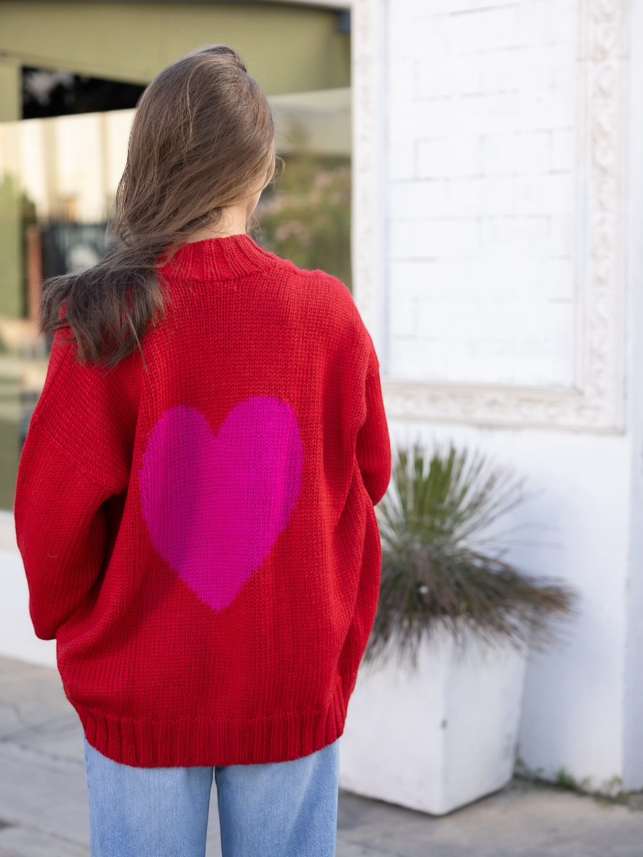Red alpaca cardigan featuring an oversized pink heart motif on the back, shown here on a model in an outdoor urban setting.