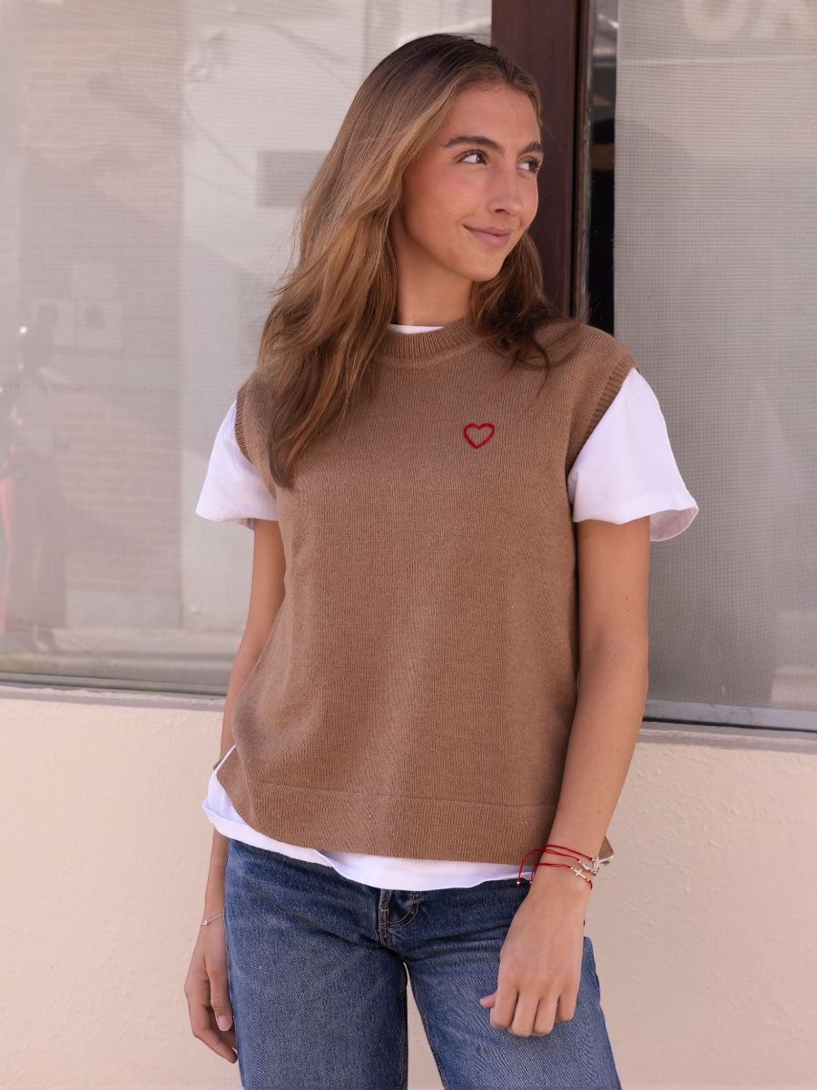 Alpaca sweater vest in a camel color with a red heart detail on model with a wall background