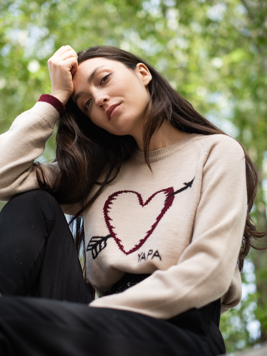 beige alpaca sweater featuring a heart with an arrow design on the chest on a model in a park background