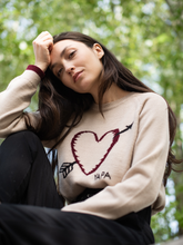 Load image into Gallery viewer, beige alpaca sweater featuring a heart with an arrow design on the chest on a model in a park background
