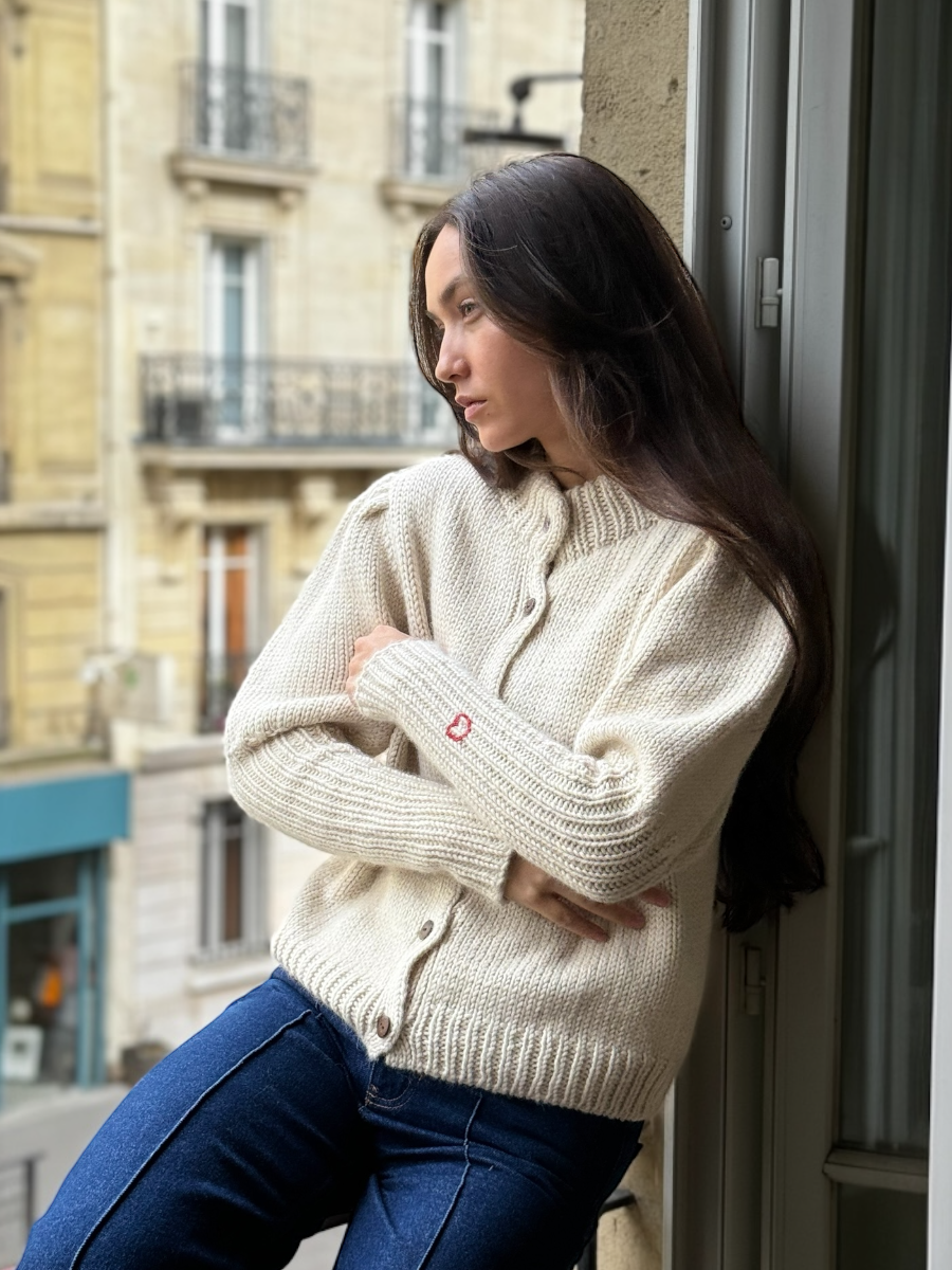 White alpaca cardigan featuring puffed sleeves, a ribbed high neckline, and a small red embroidered heart on the cuff, shown here on a model in an indoor setting.