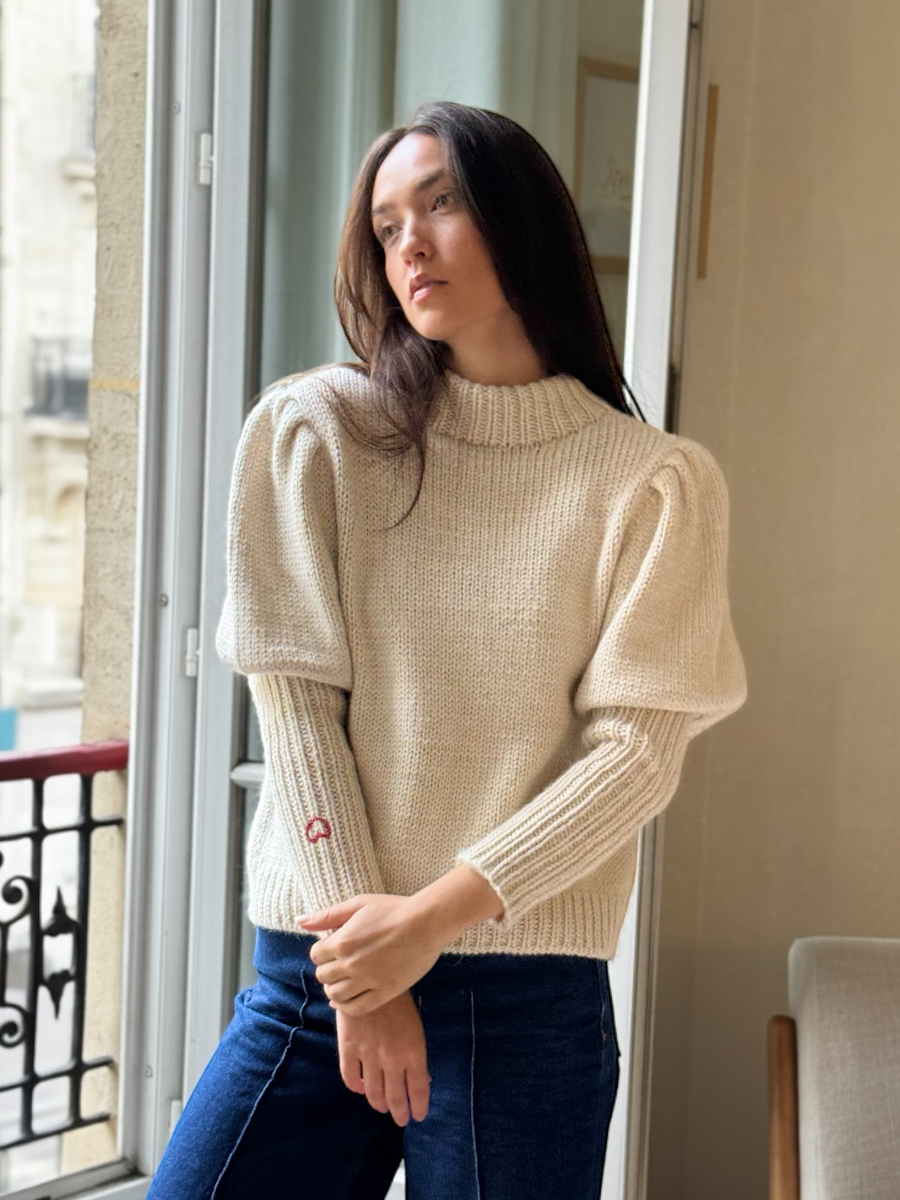 White alpaca cardigan featuring puffed sleeves, a ribbed high neckline, and a small red embroidered heart on the cuff, shown here on a model in an indoor setting.