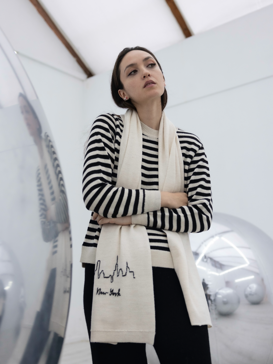 White alpaca scarf featuring a hand-embroidered black New York City skyline motif on the front, shown here on a model in an art gallery.