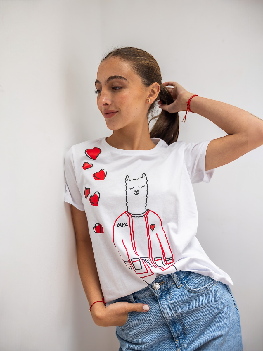 White cotton t-shirt featuring a playful embroidered alpaca in a red jacket with scattered red heart motifs and the word “YAPA,” shown here on a model in a casual indoor setting.