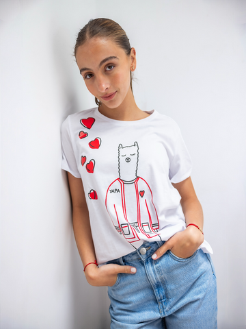 White cotton t-shirt featuring a playful embroidered alpaca in a red jacket with scattered red heart motifs and the word “YAPA,” shown here on a model in a casual indoor setting.