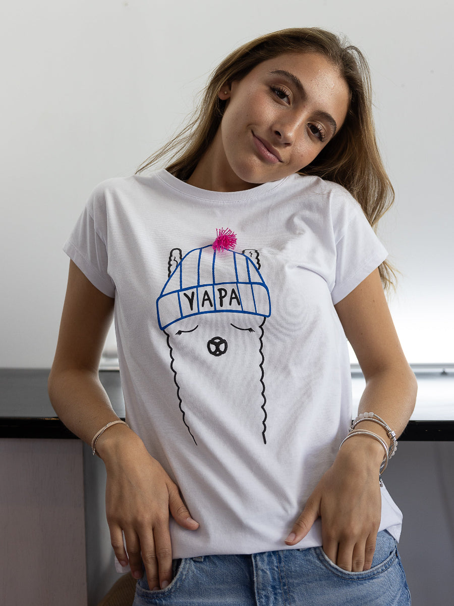 White cotton t-shirt featuring a playful embroidered alpaca motif wearing a blue beanie with a pink tassel and the word “YAPA” on the front, worn by a model.