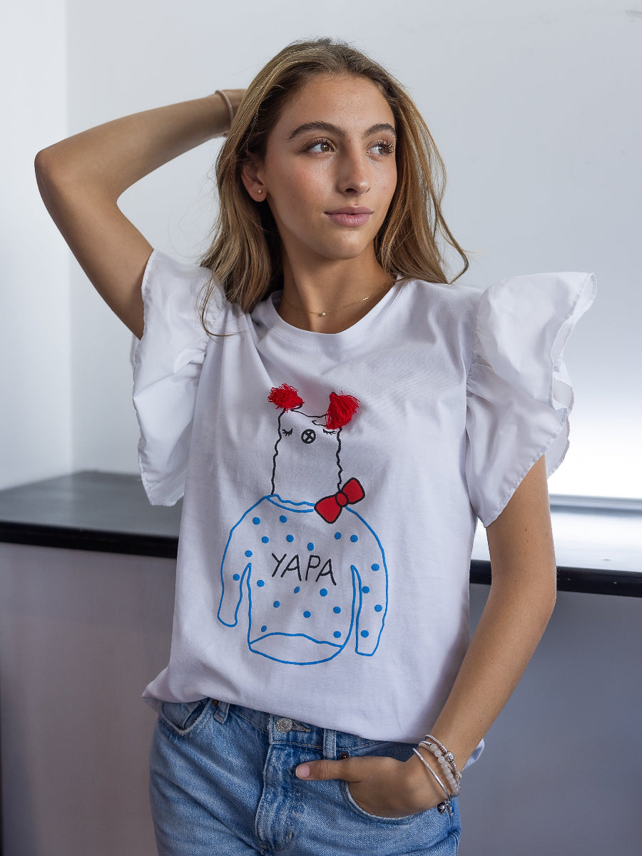 White cotton t-shirt featuring a playful embroidered alpaca motif with red tassel details, a red bow, and the word “YAPA” on the front and a ruffled design on the shoulders, shown here on a model