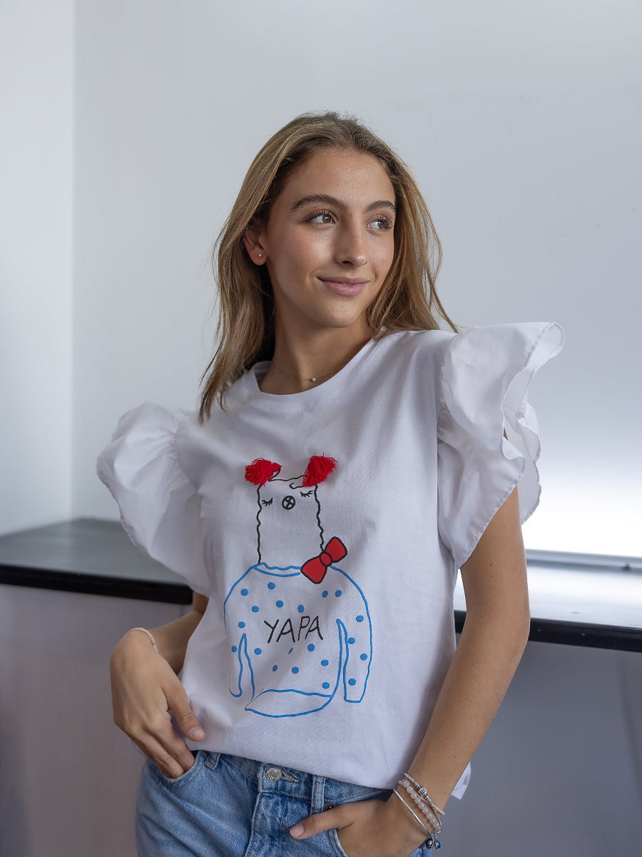 White cotton t-shirt featuring a playful embroidered alpaca motif with red tassel details, a red bow, and the word “YAPA” on the front and a ruffled design on the shoulders, shown here on a model