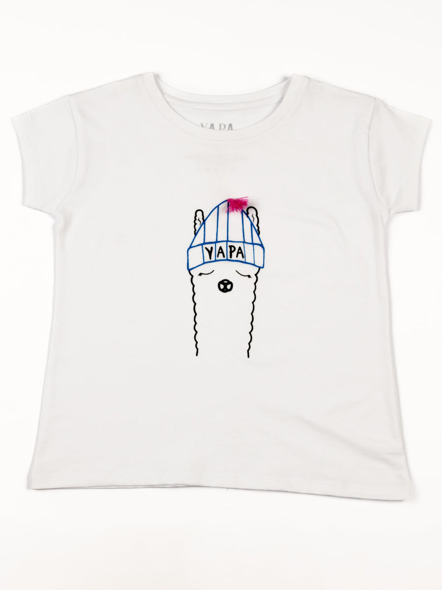 White cotton t-shirt featuring a playful embroidered alpaca motif wearing a blue beanie with a pink tassel and the word “YAPA” on the front