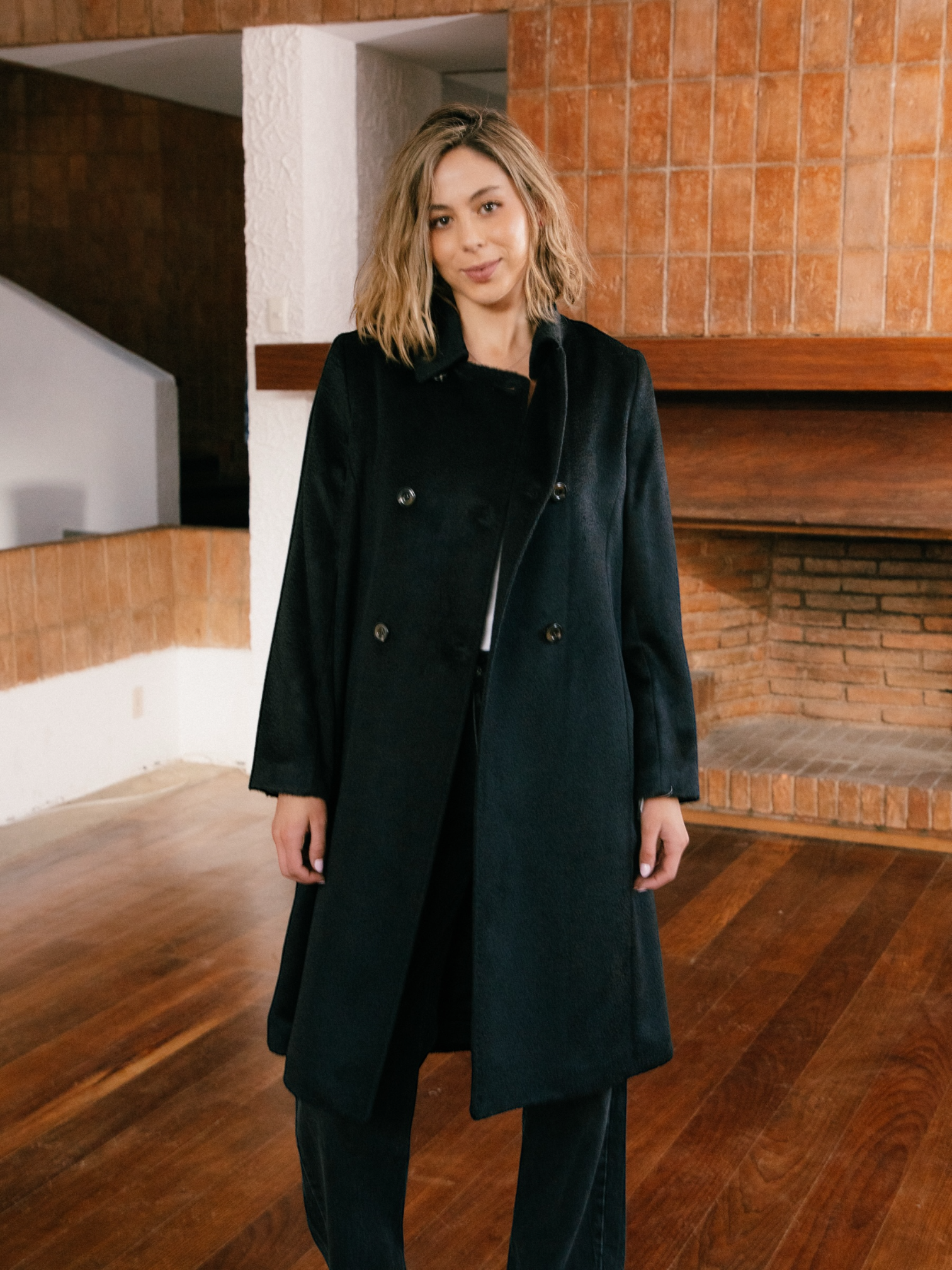 Woman wearing a black alpaca coat standing in a modern architectural setting with wooden floors and walls.