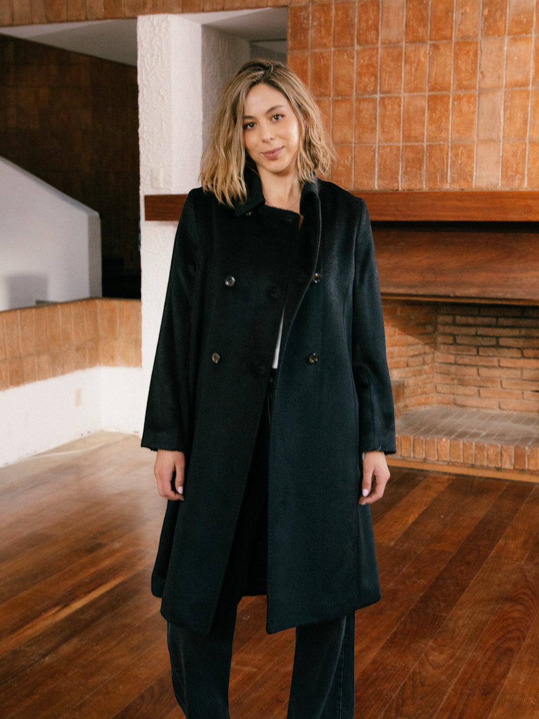 Woman wearing a black alpaca coat standing in a modern architectural setting with wooden floors and walls.