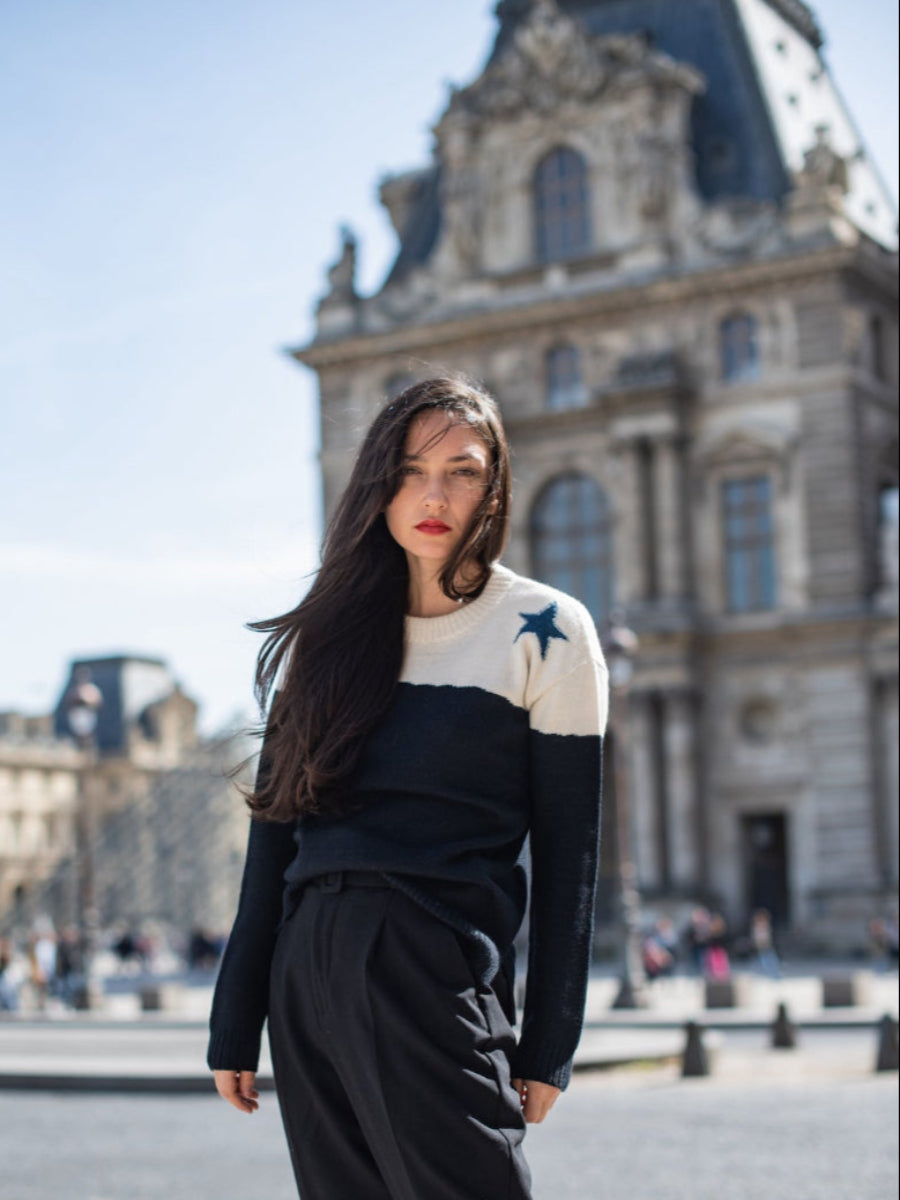 A black and white alpaca sweater with a blue star detail on the shoulder, worn by a model in an outdoor city setting