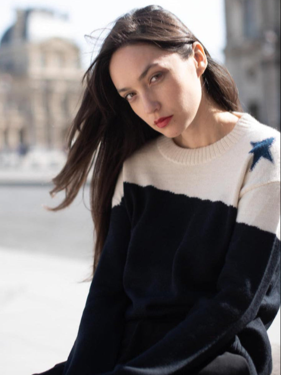 A black and cream alpaca sweater with a blue star detail on the shoulder, worn by a model in an outdoor city setting